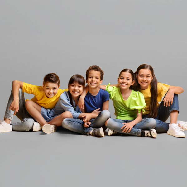 Happy carefree childhood concept. Lovely cheerful diverse kids multiracial preteen school aged boys and girls friends sitting on floor, embracing and smiling at camera on yellow background